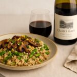 A plate of African-inspired mushroom risotto with spring vegetables, paired with a glass and bottle of Quails' Gate Pinot Noir on a light neutral background.