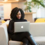 A confident Black woman in a sunny room with a laptop on a sofa.