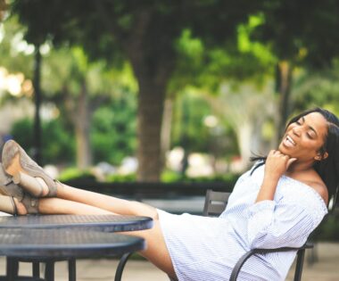 Black woman relaxing outdoors at a café table with her feet up, smiling joyfully in soft natural light