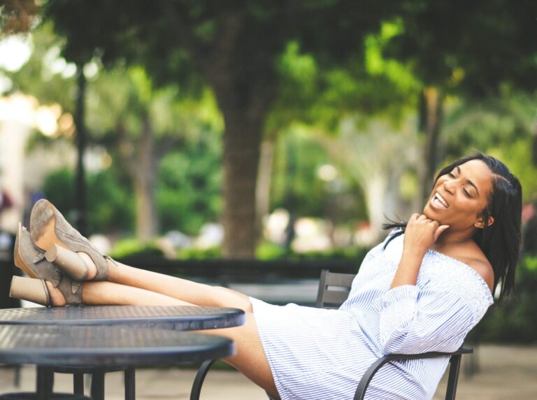 Black woman relaxing outdoors at a café table with her feet up, smiling joyfully in soft natural light