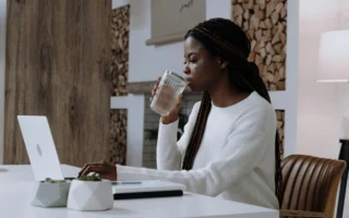 Black woman with long braids drinking water while working on a laptop at a modern white desk in a cozy home office.