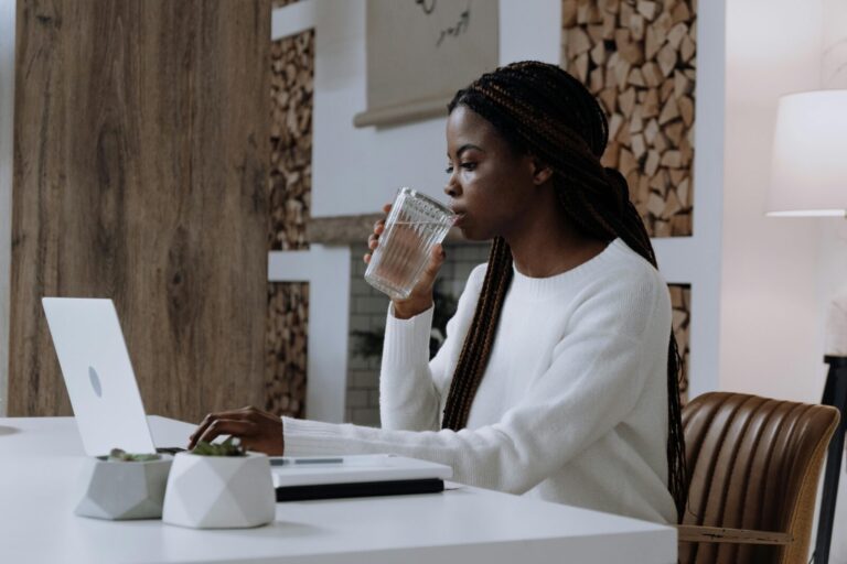 Black woman with long braids drinking water while working on a laptop at a modern white desk in a cozy home office.