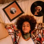 A relaxed Black woman lying on a bed surrounded by vinyl records, enjoying a moment of nostalgia and peace
