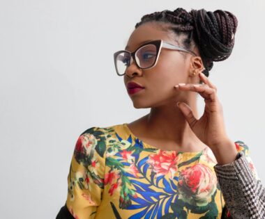 Black woman in a vibrant yellow floral dress and oversized glasses, gazing thoughtfully to the side against a clean white backdrop.