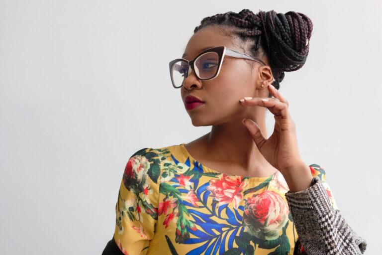Black woman in a vibrant yellow floral dress and oversized glasses, gazing thoughtfully to the side against a clean white backdrop.