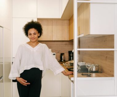 A young Black woman with natural curly hair stands confidently in a bright, modern kitchen featuring minimalist decor, soft wood accents, and white cabinetry. She wears a casual white blouse paired with dark pants, creating a chic, relaxed atmosphere.