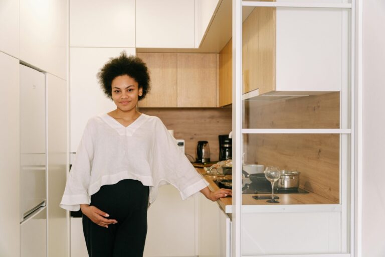 A young Black woman with natural curly hair stands confidently in a bright, modern kitchen featuring minimalist decor, soft wood accents, and white cabinetry. She wears a casual white blouse paired with dark pants, creating a chic, relaxed atmosphere.
