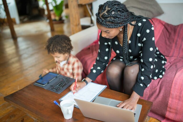 "A Black mother multitasking, working on a laptop and paperwork at home, while her child uses a tablet beside her, in a cozy, bright living room setting