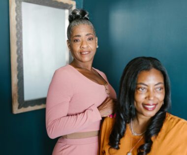 Two joyful middle-aged Black women standing close together against a blue background, one with gray hair in a bun and the other with long black hair, smiling warmly.