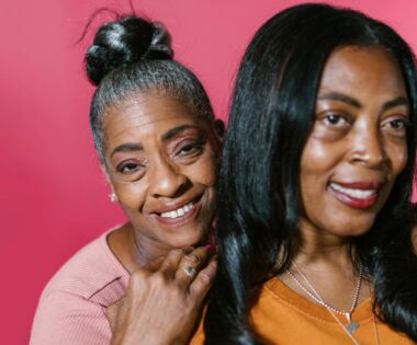 Two joyful middle-aged Black women standing close together against a pink background, one with gray hair in a bun and the other with long black hair, smiling warmly.