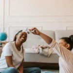 Alt text: A joyful moment between a Black mother and her daughter as they playfully interact at home, smiling and bonding in a warmly lit, cozy living room.