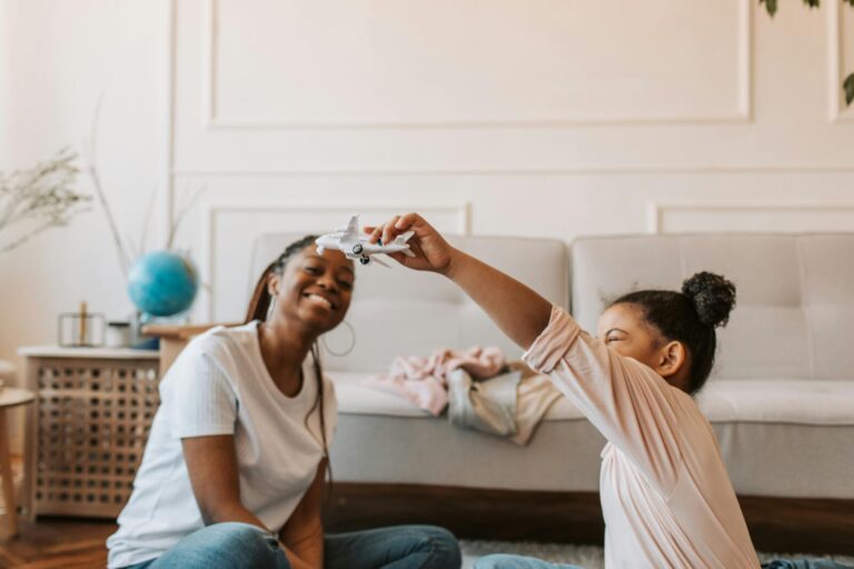 Alt text: A joyful moment between a Black mother and her daughter as they playfully interact at home, smiling and bonding in a warmly lit, cozy living room.