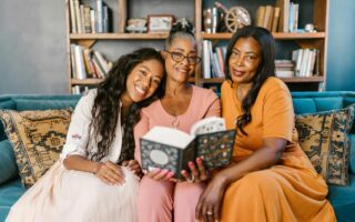 Three Black women of different generations sitting on a cozy couch, smiling and sharing a book together in a warmly lit living room”