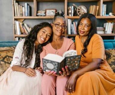 Three Black women of different generations sitting on a cozy couch, smiling and sharing a book together in a warmly lit living room”