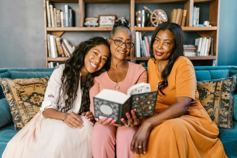 Three Black women of different generations sitting on a cozy couch, smiling and sharing a book together in a warmly lit living room”